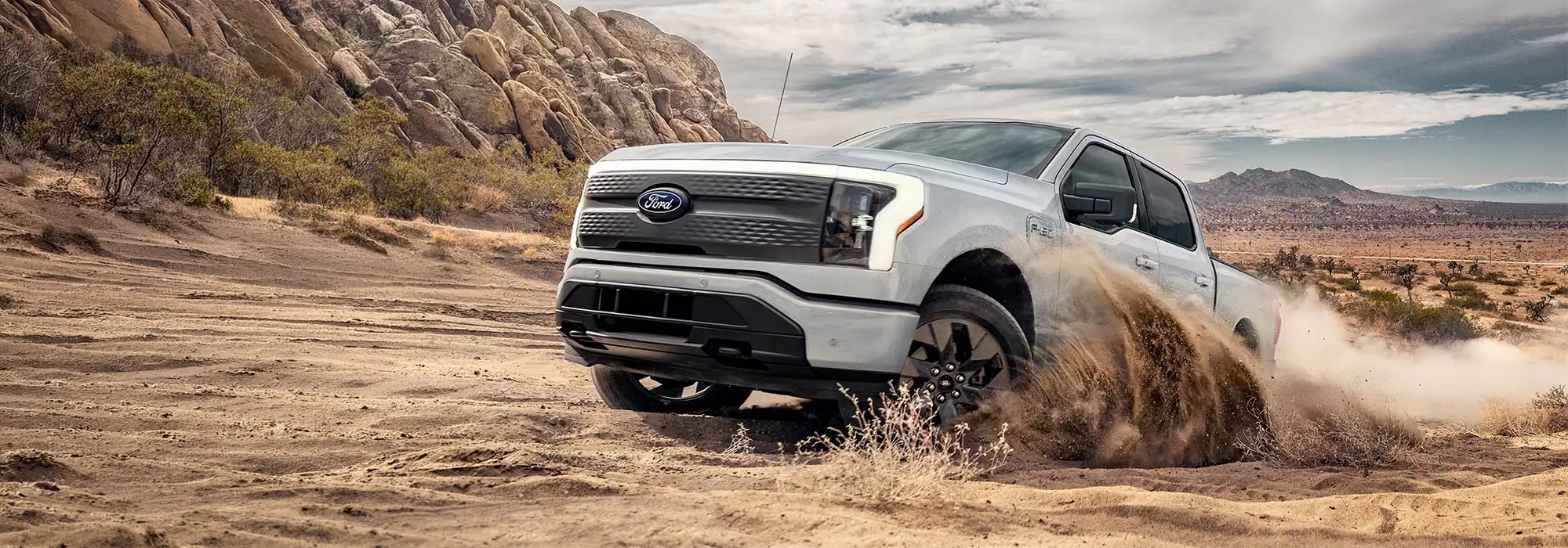 Silver Ford F-150 Lightning electric pickup truck driving through desert sand and kicking up dust.